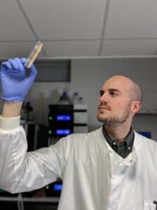 Man wearing lab coat holding a Petri dish up to the light. 